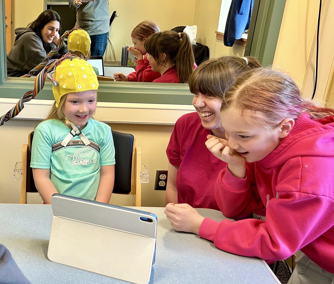 5-year-old girl wearing an EEG cap and looking at an ipad with older sister and two researchers.